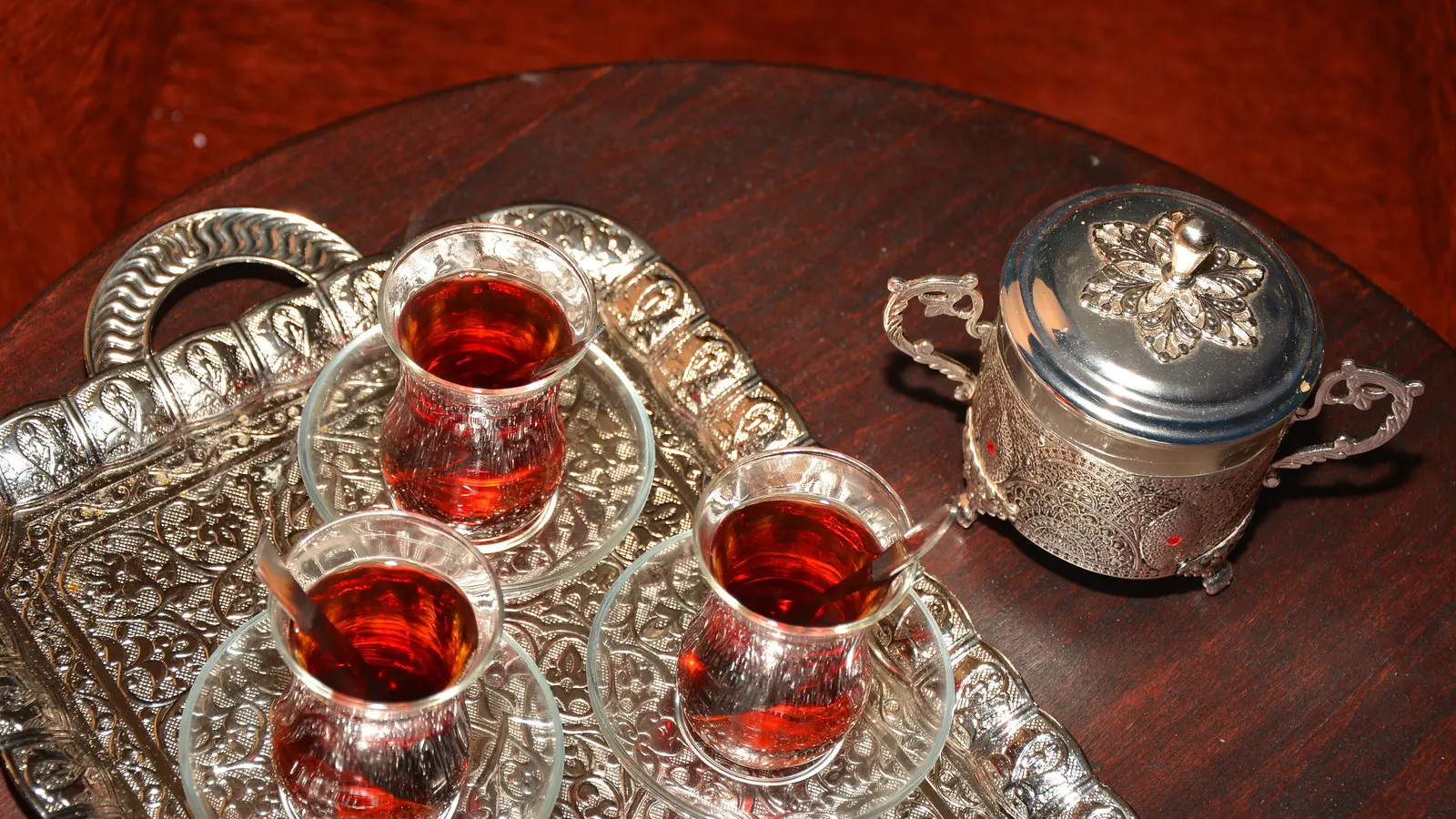 Gümüş tepside üç ince belli çay bardağı ve kapaklı şekerlik; bardaklar dolu, ahşap masa üzerinde duruyor.