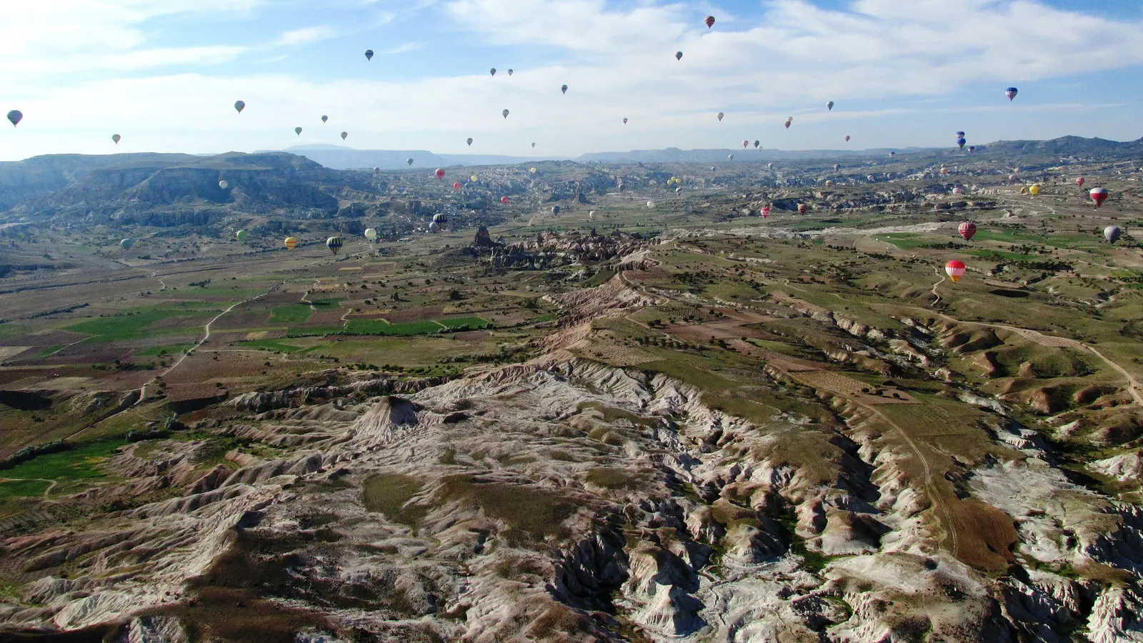 Kapadokya vadilerinin üzerinde aynı sabah havalanmış onlarca sıcak hava balonu; arkada geniş plato ve açık gökyüzü.