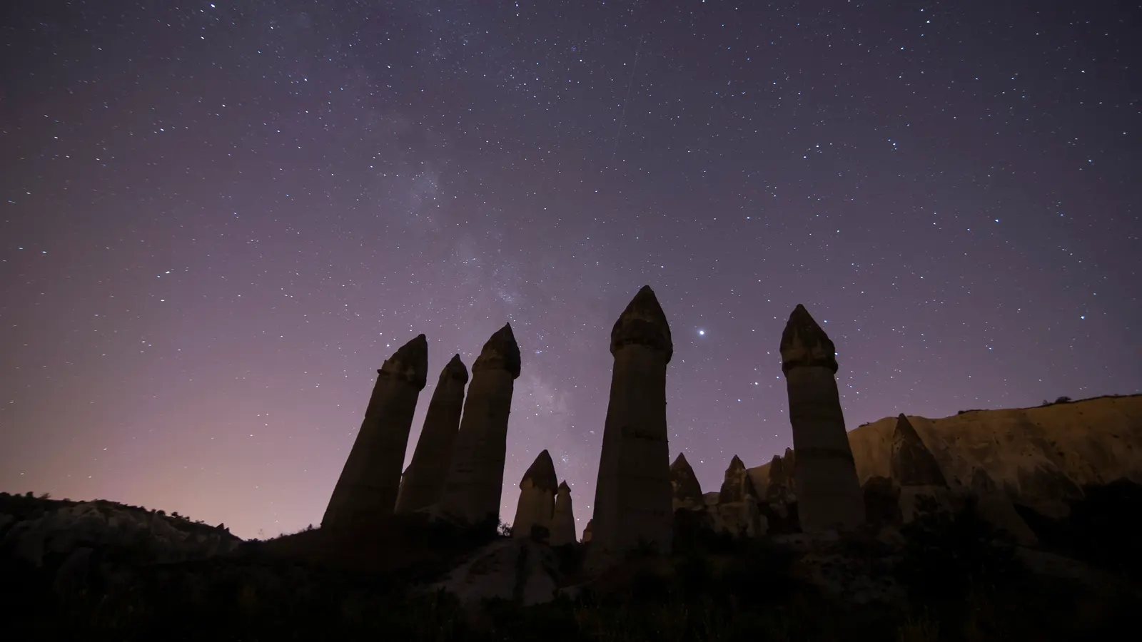 Kapadokya’da gece: yıldızlarla dolu gökyüzünde soluk bir Samanyolu bandı ve siluet hâlinde yükselen peri bacaları; ufukta hafif ışık parıltısı.