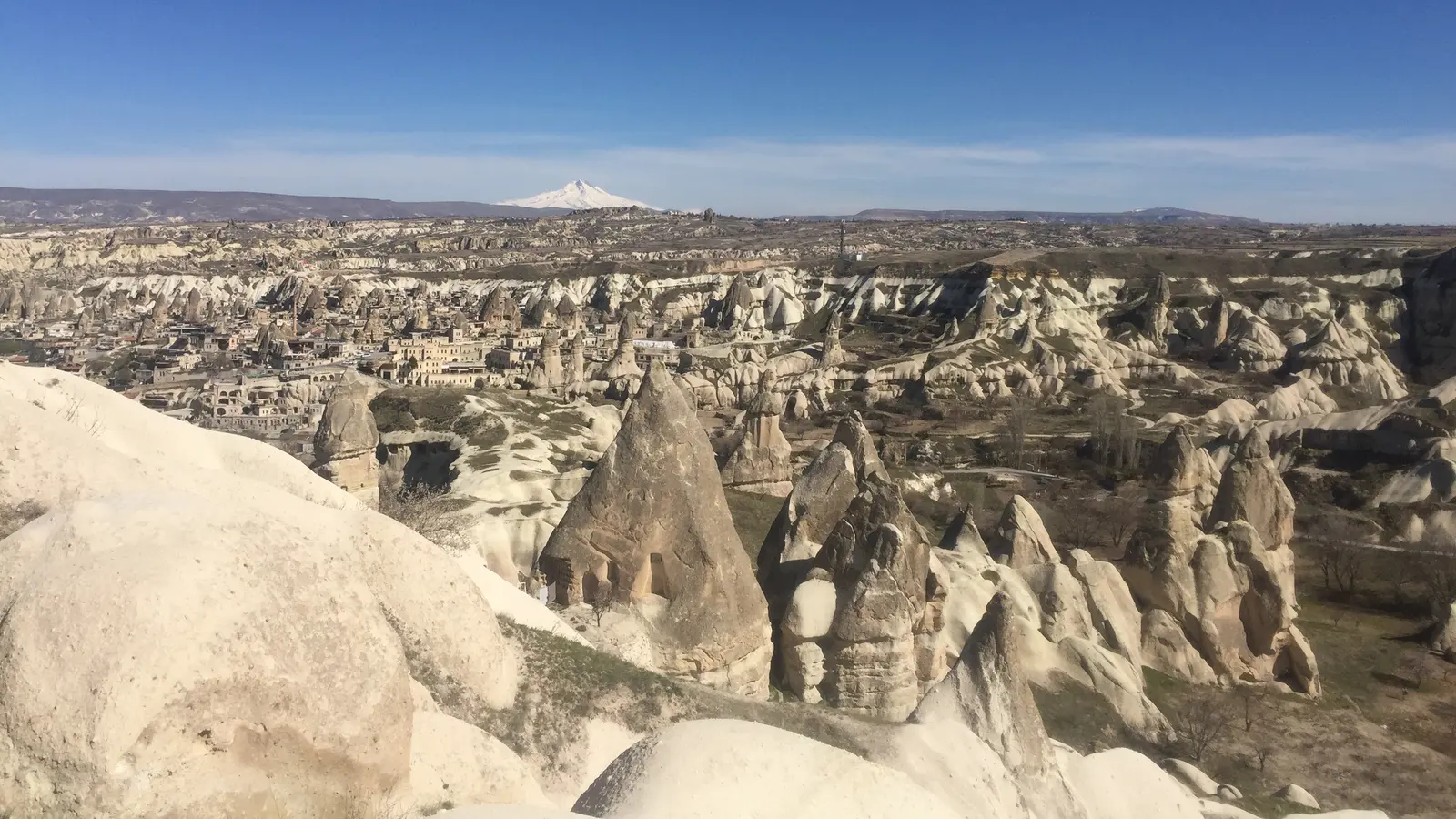 Göreme'nin bir seyir noktasından panoramik görünümü; önde peri bacaları, uzakta karlı bir dağ ve açık mavi gökyüzü.